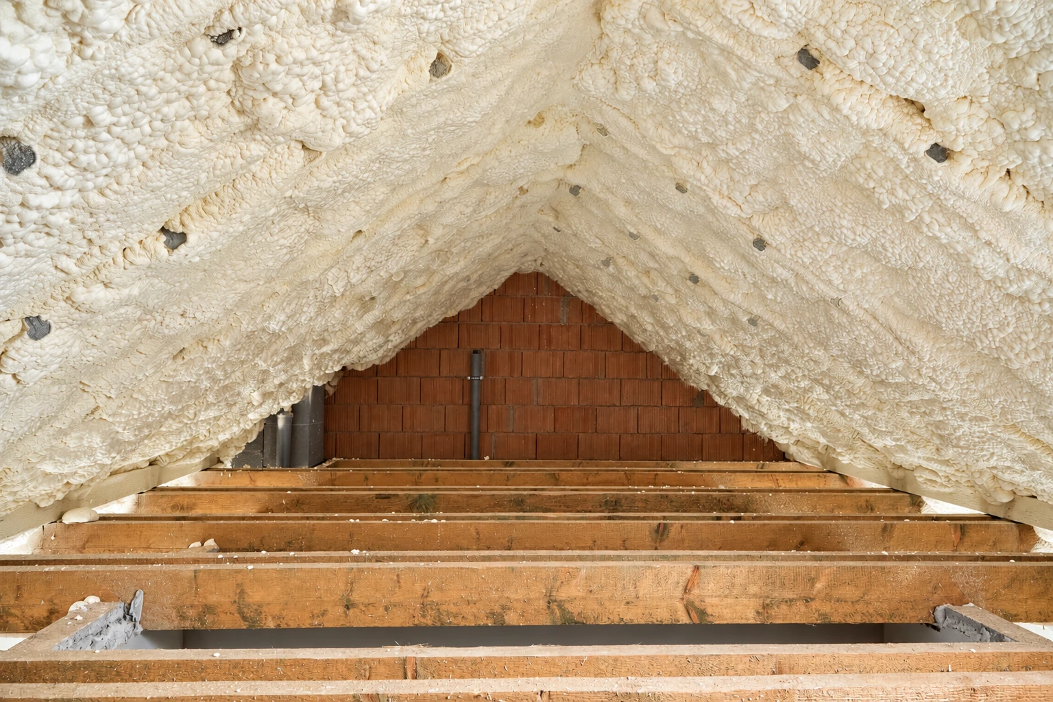 Attic roof slopes covered in thick white spray foam above exposed floor joists and a brick gable wall.