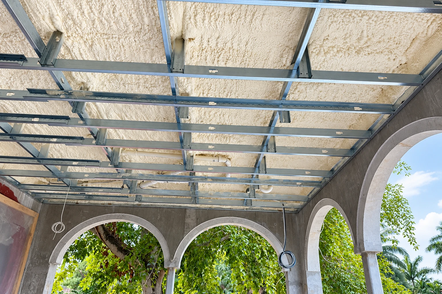 Spray foam covers a commercial building ceiling above a grid of exposed metal framing and arched openings.