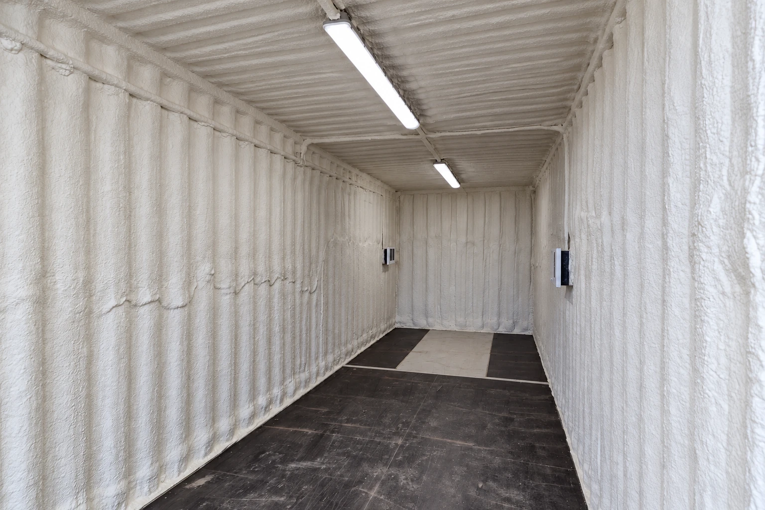 A narrow commercial interior is coated with white spray foam insulation across its walls and ceiling.