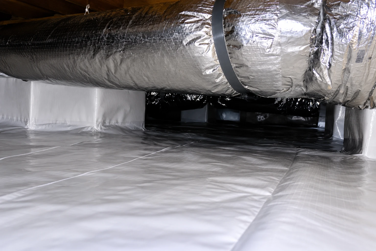 Reflective insulated ductwork runs through a crawl space above a floor covered with white vapor barrier.