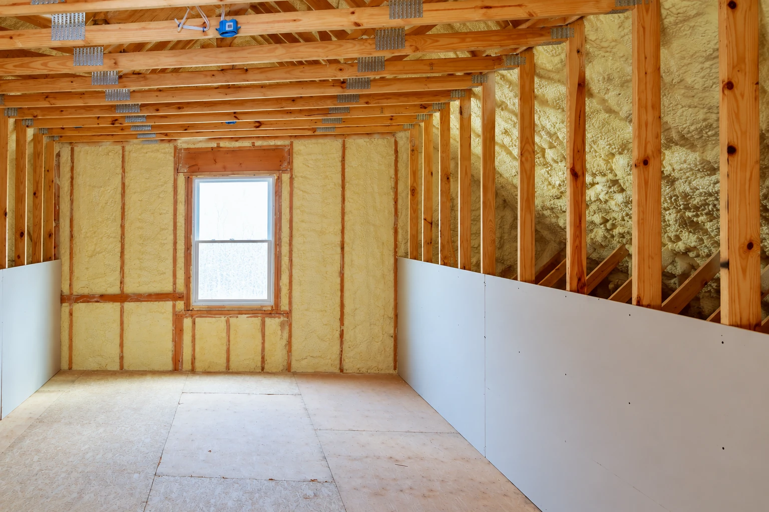 A framed attic room shows yellow spray foam insulation in the walls and roof, with drywall panels partly installed.