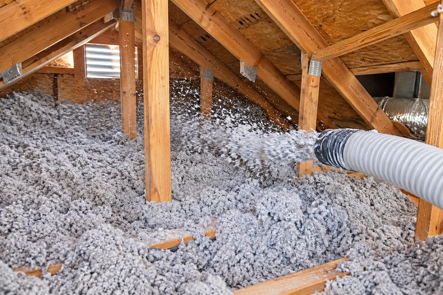 Blown-in insulation is being sprayed from a flexible hose across the floor of a wooden attic.