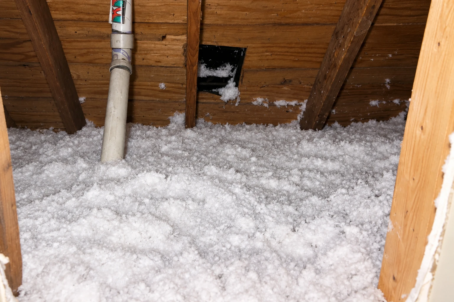 Loose-fill insulation covers an attic floor beneath a wooden roof, surrounding a vertical pipe.