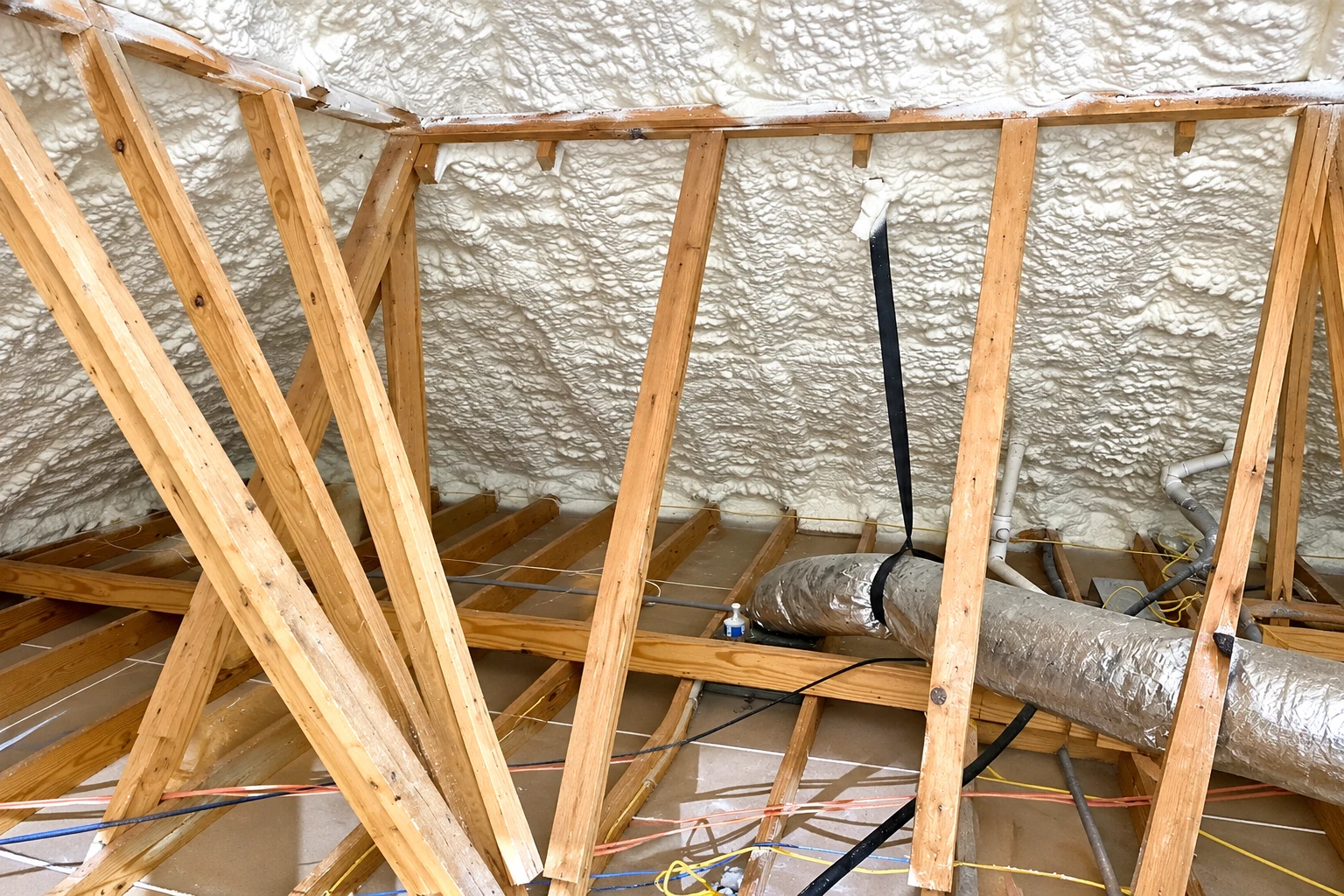 Spray foam insulation coats the roof and walls of a timber attic, with exposed rafters, ductwork, and wiring.