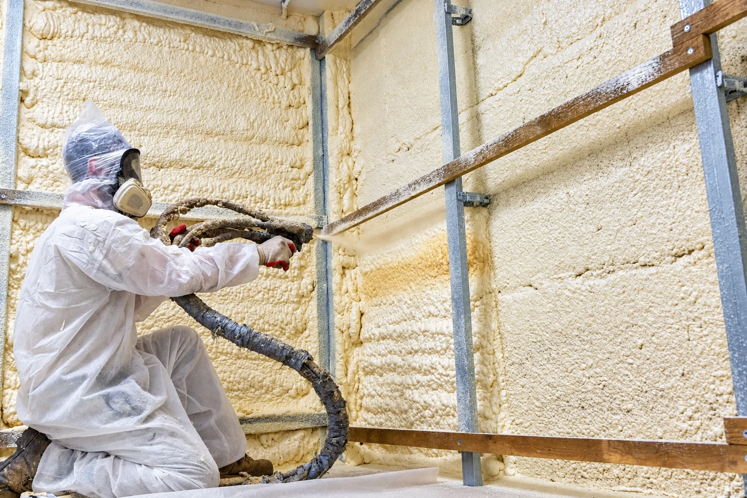A worker in protective coveralls sprays foam insulation onto a framed wall, covering it in thick cream-colored foam.